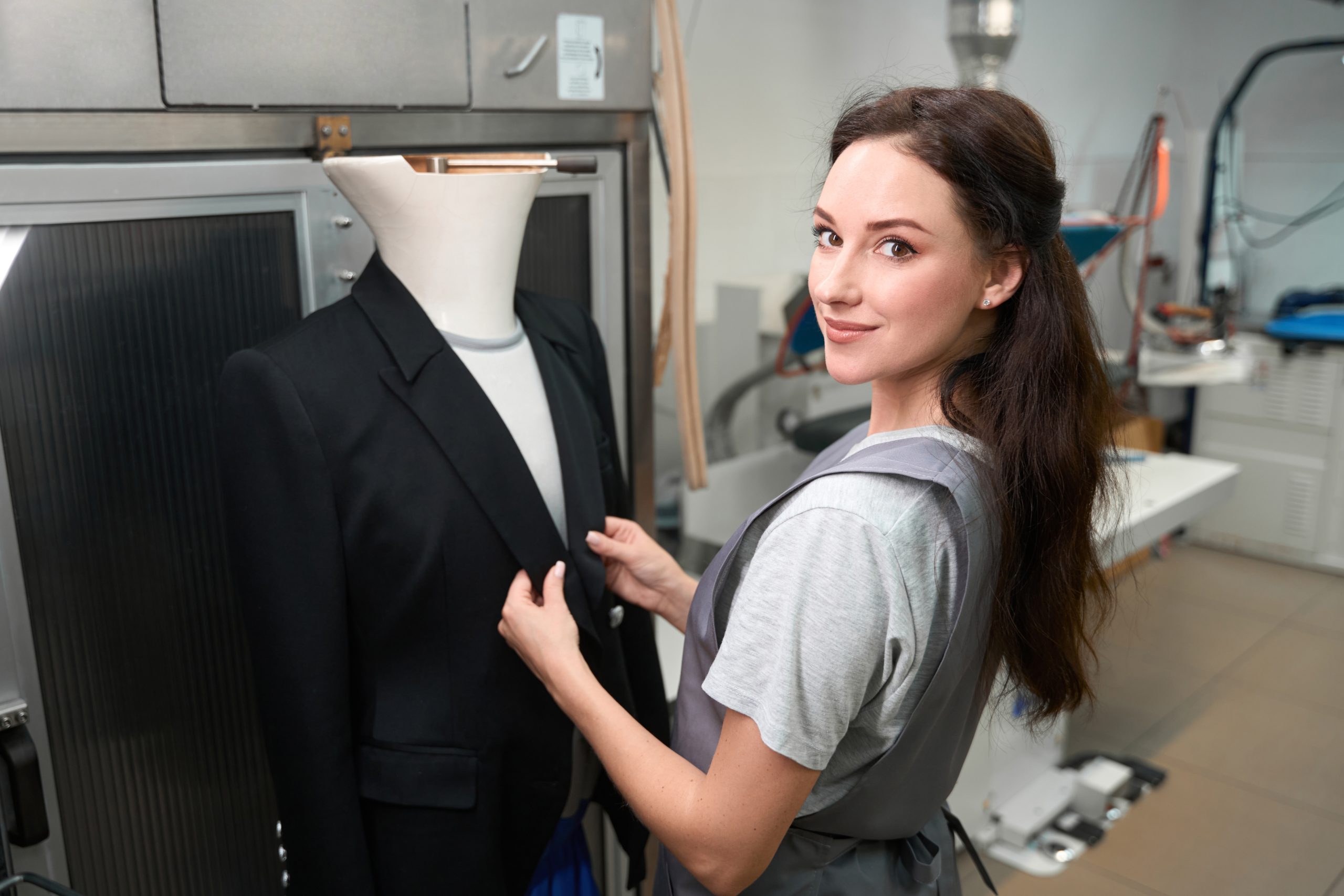 Smiling high-qualified woman laundry office worker adjusting jacket on mannequin after dry-cleaning, post-treatment